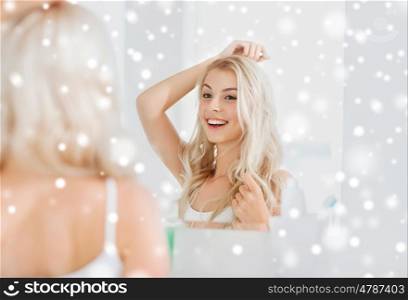 beauty, hygiene, hairstyle, morning and people concept - smiling young woman looking to mirror at home bathroom over snow