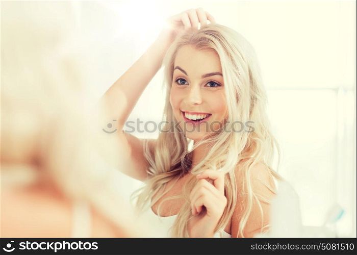 beauty, hygiene, hairstyle, morning and people concept - smiling young woman looking to mirror at home bathroom. happy young woman looking to mirror at bathroom