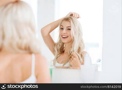 beauty, hygiene, hairstyle, morning and people concept - smiling young woman looking to mirror at home bathroom