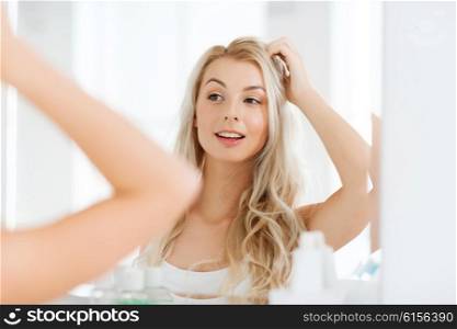 beauty, hygiene, hairstyle, morning and people concept - smiling young woman looking to mirror at home bathroom
