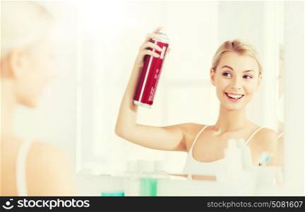 beauty, hygiene, hairstyle, morning and people concept - smiling young woman with hairspray styling her hair and looking to mirror at home bathroom. woman with hairspray styling her hair at bathroom
