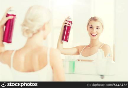 beauty, hygiene, hairstyle, morning and people concept - smiling young woman with hairspray styling her hair and looking to mirror at home bathroom. woman with hairspray styling her hair at bathroom