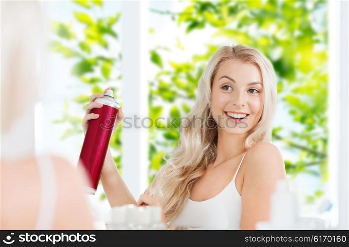 beauty, hygiene, hairstyle, morning and people concept - smiling young woman with hairspray styling her hair and looking to mirror at home bathroom over green natural background
