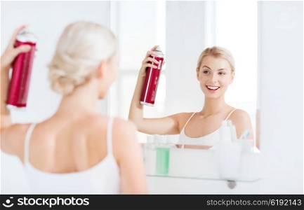 beauty, hygiene, hairstyle, morning and people concept - smiling young woman with hairspray styling her hair and looking to mirror at home bathroom
