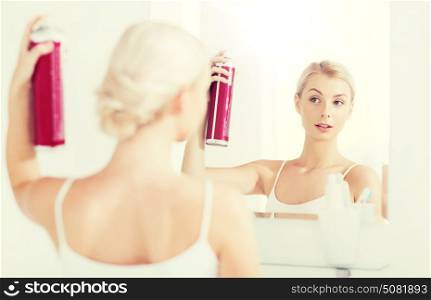beauty, hygiene, hairstyle, morning and people concept - young woman with hairspray styling her hair and looking to mirror at home bathroom. woman with hairspray styling her hair at bathroom