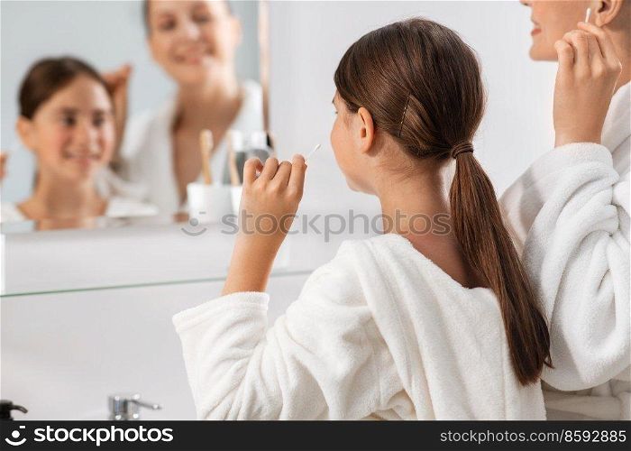 beauty, hygiene, morning and people concept - happy smiling mother and daughter cleaning ears with cotton swabs and looking to mirror at bathroom. mother and daughter cleaning ears with cotton swab