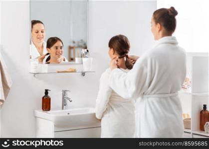 beauty, hygiene, morning and people concept - happy smiling mother and daughter looking to mirror and braiding hair at bathroom. mother and daughter braiding hair at bathroom