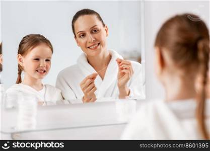 beauty, hygiene, morning and people concept - happy smiling mother and little daughter with dental floss cleaning teeth and looking to mirror at bathroom. mother and daughter with floss cleaning teeth