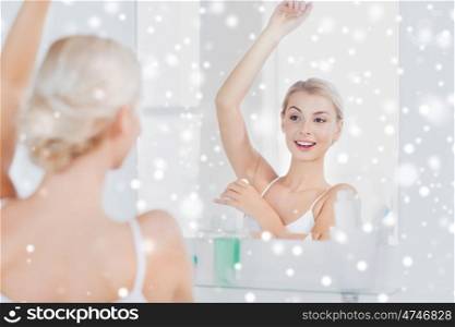 beauty, hygiene, morning and people concept - smiling young woman applying antiperspirant or stick deodorant and looking to mirror at home bathroom over snow