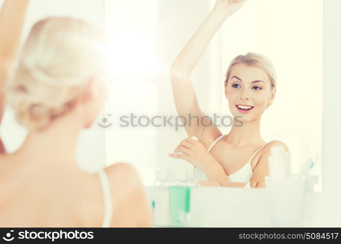 beauty, hygiene, morning and people concept - smiling young woman applying antiperspirant or stick deodorant and looking to mirror at home bathroom. woman with antiperspirant deodorant at bathroom. woman with antiperspirant deodorant at bathroom