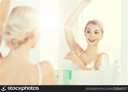 beauty, hygiene, morning and people concept - smiling young woman applying antiperspirant or stick deodorant and looking to mirror at home bathroom. woman with antiperspirant deodorant at bathroom. woman with antiperspirant deodorant at bathroom