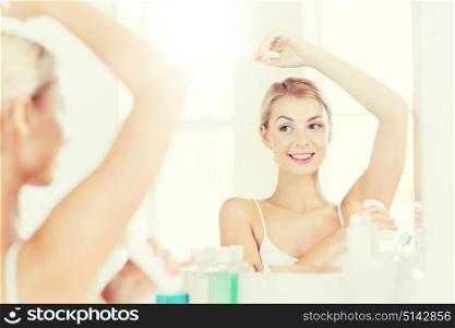 beauty, hygiene, morning and people concept - smiling young woman applying antiperspirant or stick deodorant and looking to mirror at home bathroom. woman with antiperspirant deodorant at bathroom