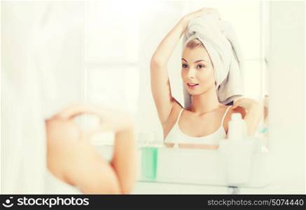 beauty, hygiene, morning and people concept - smiling young woman with head wrapper in towel looking to mirror at home bathroom. happy young woman with towel at mirror in bathroom