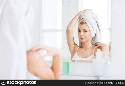 beauty, hygiene, morning and people concept - smiling young woman with head wrapper in towel looking to mirror at home bathroom