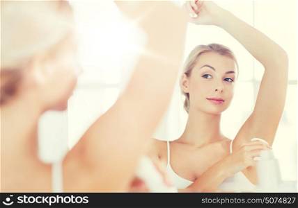 beauty, hygiene, morning and people concept - young woman applying antiperspirant or stick deodorant and looking to mirror at home bathroom. woman with antiperspirant deodorant at bathroom