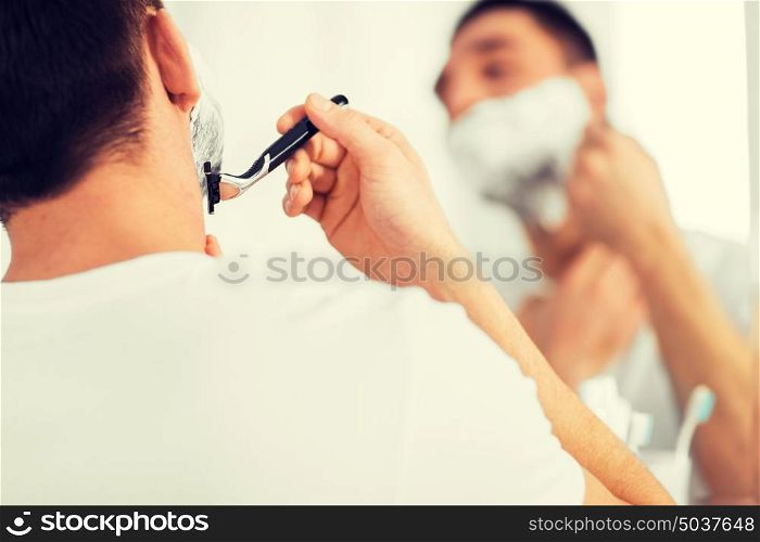 beauty, hygiene, shaving, grooming and people concept - close up of young man looking to mirror and shaving beard with manual razor blade at home bathroom. close up of man shaving beard with razor blade