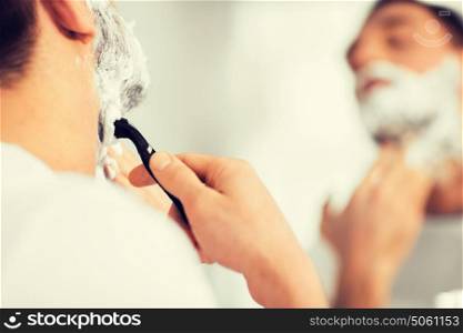 beauty, hygiene, shaving, grooming and people concept - close up of young man looking to mirror and shaving beard with manual razor blade at home bathroom. close up of man shaving beard with razor blade