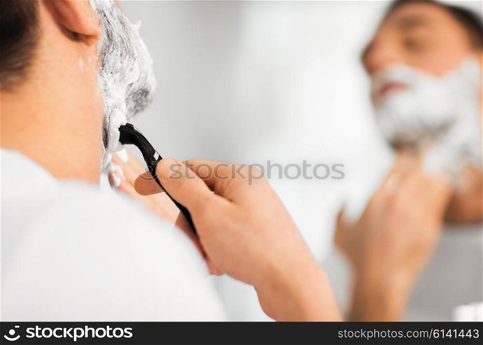 beauty, hygiene, shaving, grooming and people concept - close up of young man looking to mirror and shaving beard with manual razor blade at home bathroom