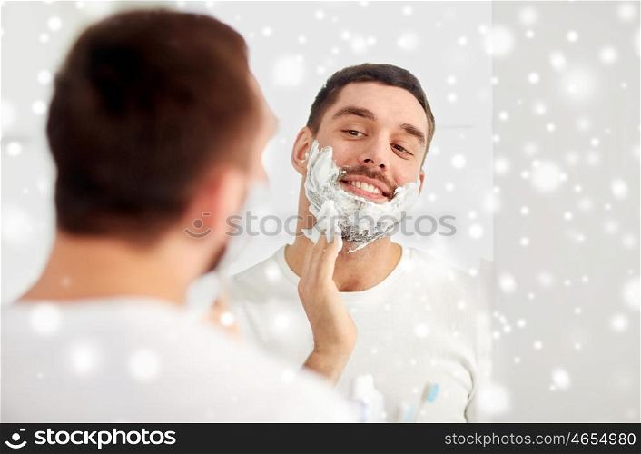 beauty, hygiene, shaving, grooming and people concept - smiling young man looking to mirror and applying shaving foam to face at home bathroom over snow