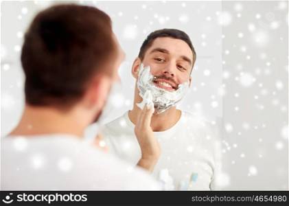 beauty, hygiene, shaving, grooming and people concept - smiling young man looking to mirror and applying shaving foam to face at home bathroom over snow