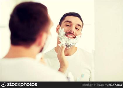 beauty, hygiene, shaving, grooming and people concept - smiling young man looking to mirror and applying shaving foam to face at home bathroom. happy man applying shaving foam at bathroom mirror