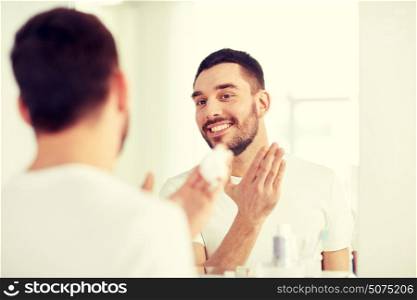 beauty, hygiene, shaving, grooming and people concept - smiling young man looking to mirror and applying shaving foam to face at home bathroom. happy man applying shaving foam at bathroom mirror
