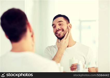 beauty, hygiene, shaving, grooming and people concept - smiling young man looking to mirror and applying aftershave at home bathroom. happy man applying aftershave at bathroom mirror. happy man applying aftershave at bathroom mirror