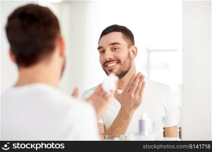beauty, hygiene, shaving, grooming and people concept - smiling young man looking to mirror and applying shaving foam to face at home bathroom