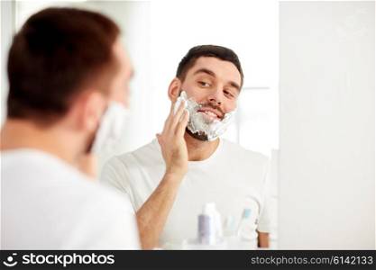beauty, hygiene, shaving, grooming and people concept - smiling young man looking to mirror and applying shaving foam to face at home bathroom