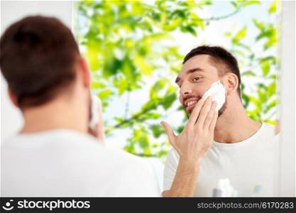 beauty, hygiene, shaving, grooming and people concept - smiling young man looking to mirror and applying shaving foam to face at home bathroom over green natural background
