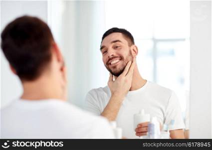 beauty, hygiene, shaving, grooming and people concept - smiling young man looking to mirror and applying aftershave at home bathroom