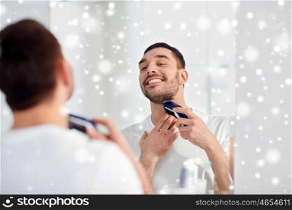 beauty, hygiene, shaving, grooming and people concept - young man looking to mirror and shaving beard with trimmer or electric shaver at home bathroom over snow