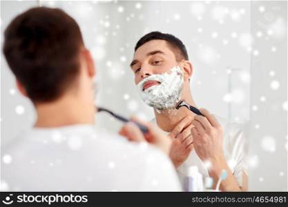 beauty, hygiene, shaving, grooming and people concept - young man looking to mirror and shaving beard with manual razor blade at home bathroom over snow