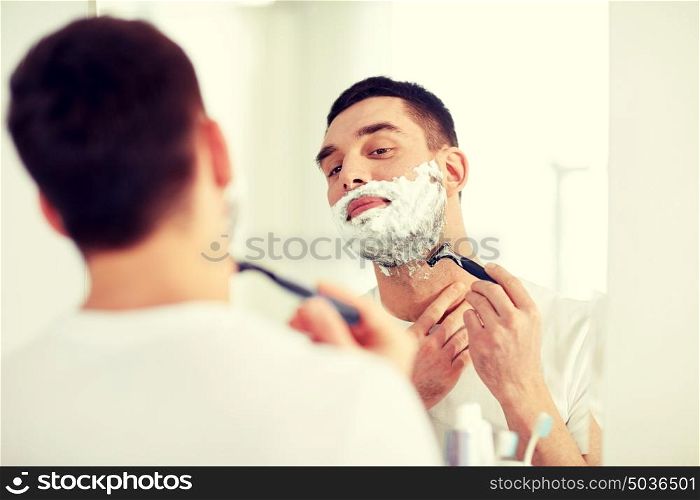 beauty, hygiene, shaving, grooming and people concept - young man looking to mirror and shaving beard with manual razor blade at home bathroom. man shaving beard with razor blade at bathroom