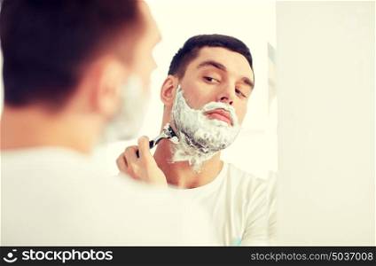 beauty, hygiene, shaving, grooming and people concept - young man looking to mirror and shaving beard with manual razor blade at home bathroom. man shaving beard with razor blade at bathroom