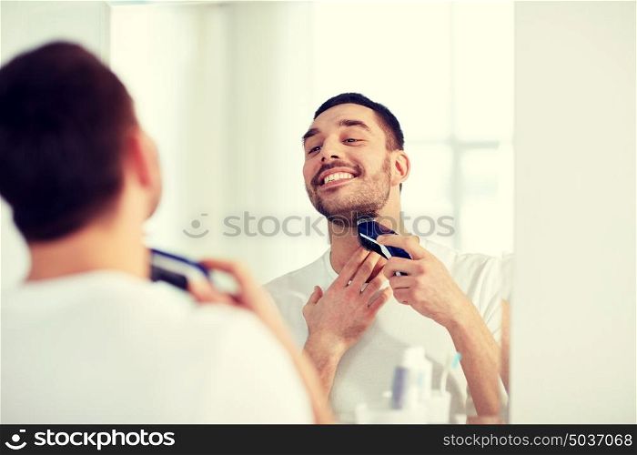 beauty, hygiene, shaving, grooming and people concept - young man looking to mirror and shaving beard with trimmer or electric shaver at home bathroom. man shaving beard with trimmer at bathroom