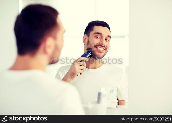 beauty, hygiene, shaving, grooming and people concept - young man looking to mirror and shaving beard with trimmer or electric shaver at home bathroom. man shaving beard with trimmer at bathroom