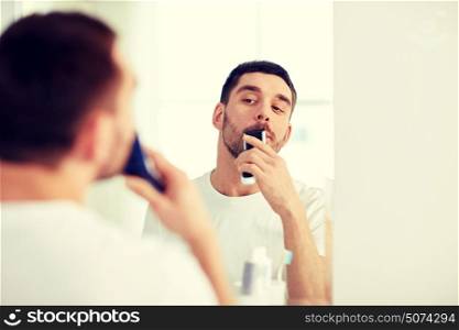beauty, hygiene, shaving, grooming and people concept - young man looking to mirror and shaving beard and mustache with trimmer or electric shaver at home bathroom. man shaving mustache with trimmer at bathroom