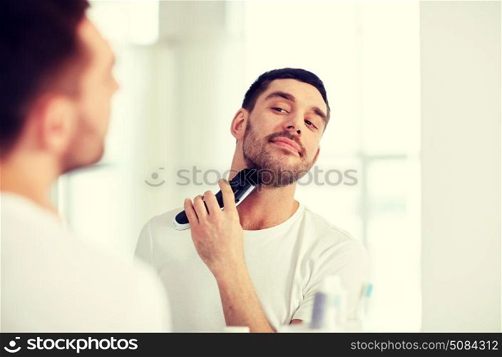 beauty, hygiene, shaving, grooming and people concept - young man looking to mirror and shaving beard with trimmer or electric shaver at home bathroom. man shaving beard with trimmer at bathroom. man shaving beard with trimmer at bathroom