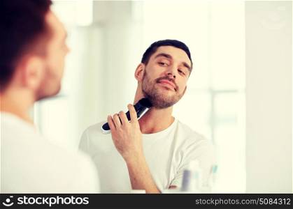 beauty, hygiene, shaving, grooming and people concept - young man looking to mirror and shaving beard with trimmer or electric shaver at home bathroom. man shaving beard with trimmer at bathroom. man shaving beard with trimmer at bathroom