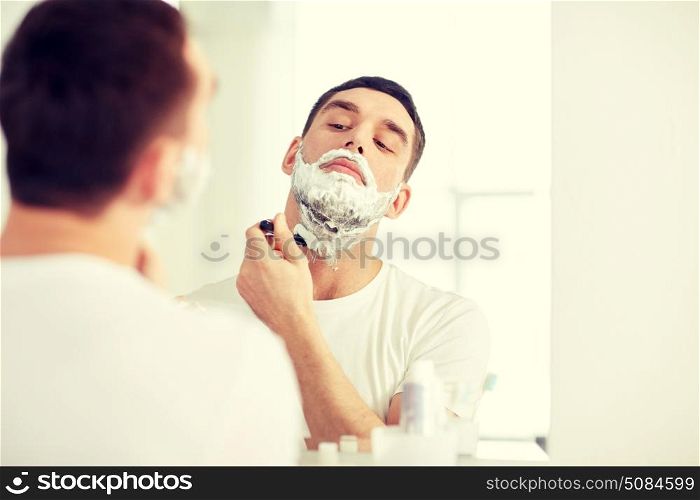 beauty, hygiene, shaving, grooming and people concept - young man looking to mirror and shaving beard with manual razor blade at home bathroom. man shaving beard with razor blade at bathroom. man shaving beard with razor blade at bathroom