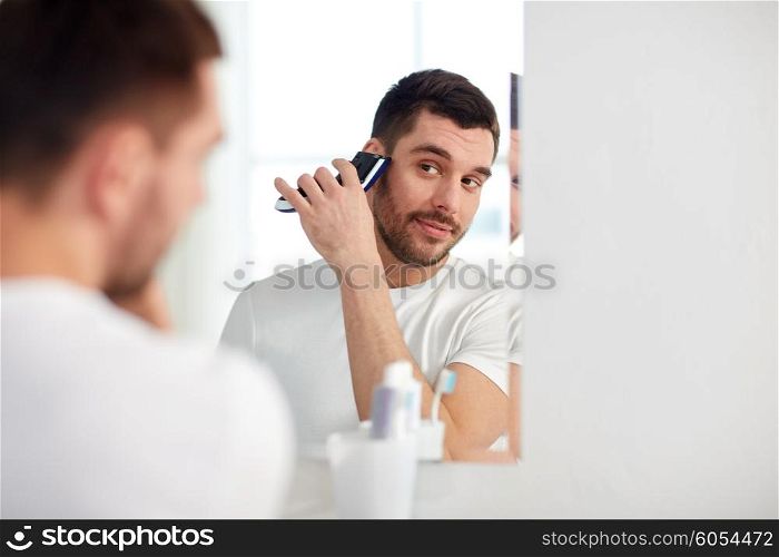 beauty, hygiene, shaving, grooming and people concept - young man looking to mirror and shaving beard with trimmer or electric shaver at home bathroom