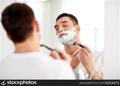 beauty, hygiene, shaving, grooming and people concept - young man looking to mirror and shaving beard with manual razor blade at home bathroom