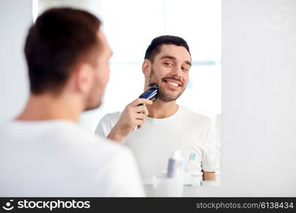 beauty, hygiene, shaving, grooming and people concept - young man looking to mirror and shaving beard with trimmer or electric shaver at home bathroom