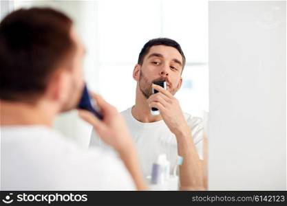 beauty, hygiene, shaving, grooming and people concept - young man looking to mirror and shaving beard and mustache with trimmer or electric shaver at home bathroom