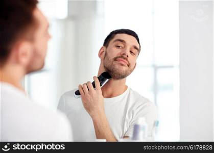 beauty, hygiene, shaving, grooming and people concept - young man looking to mirror and shaving beard with trimmer or electric shaver at home bathroom
