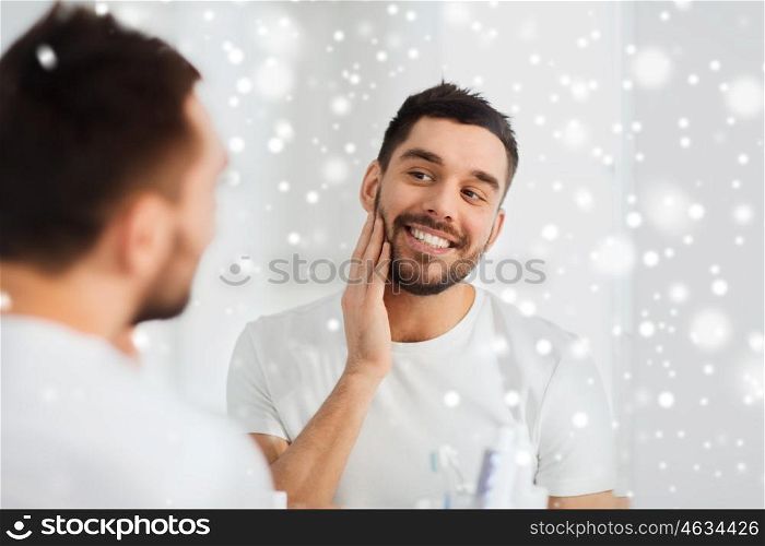 beauty, hygiene, shaving, winter and people concept - smiling young man looking to mirror at home bathroom over snow
