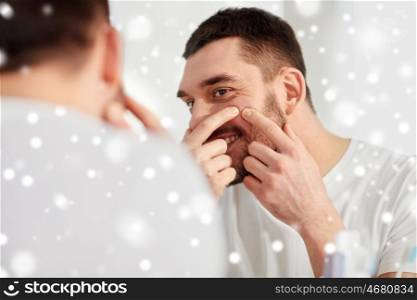 beauty, hygiene, skin problem and people concept - smiling young man looking to mirror and squeezing pimple at home bathroom over snow