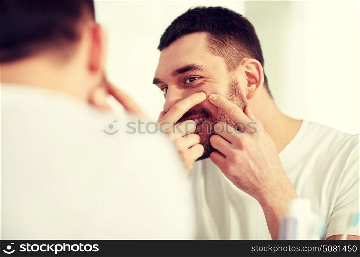 beauty, hygiene, skin problem and people concept - smiling young man looking to mirror and squeezing pimple at home bathroom. smiling man squeezing pimple at bathroom mirror