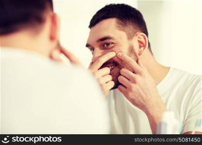 beauty, hygiene, skin problem and people concept - smiling young man looking to mirror and squeezing pimple at home bathroom. smiling man squeezing pimple at bathroom mirror